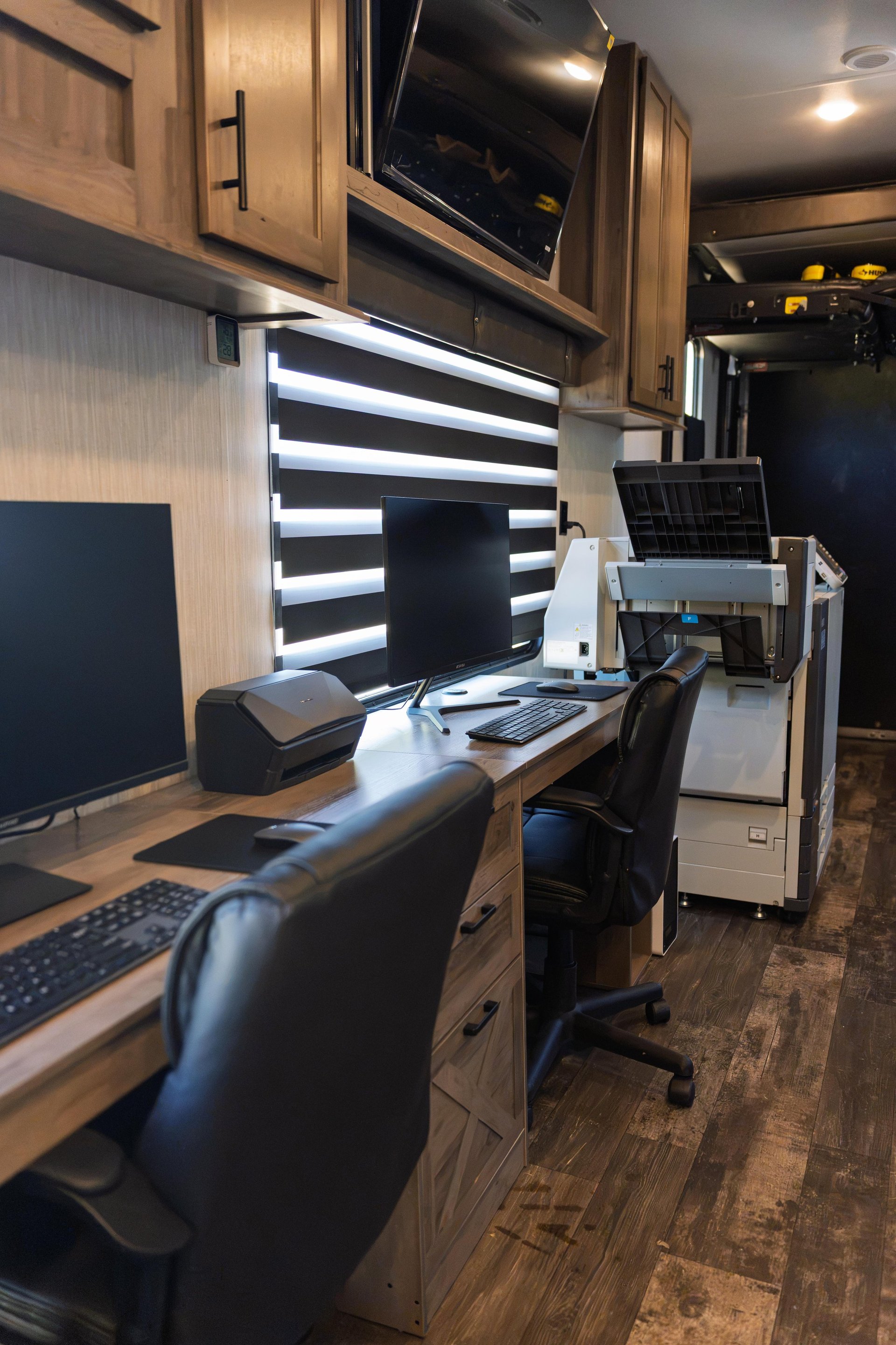Modern RV office workspace with dual monitors, keyboard, and black office chairs at wooden desk under cabinetry