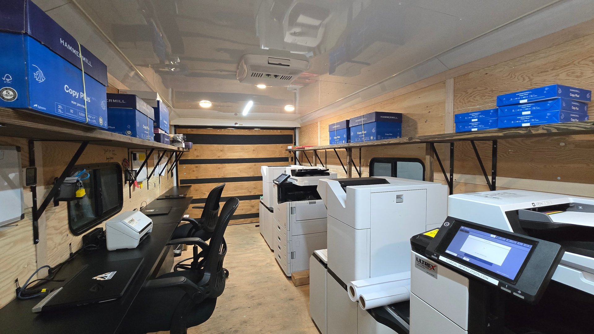Interior of a commercial print shop with multifunction printers, blue storage bins on shelves, and wooden-topped work stations