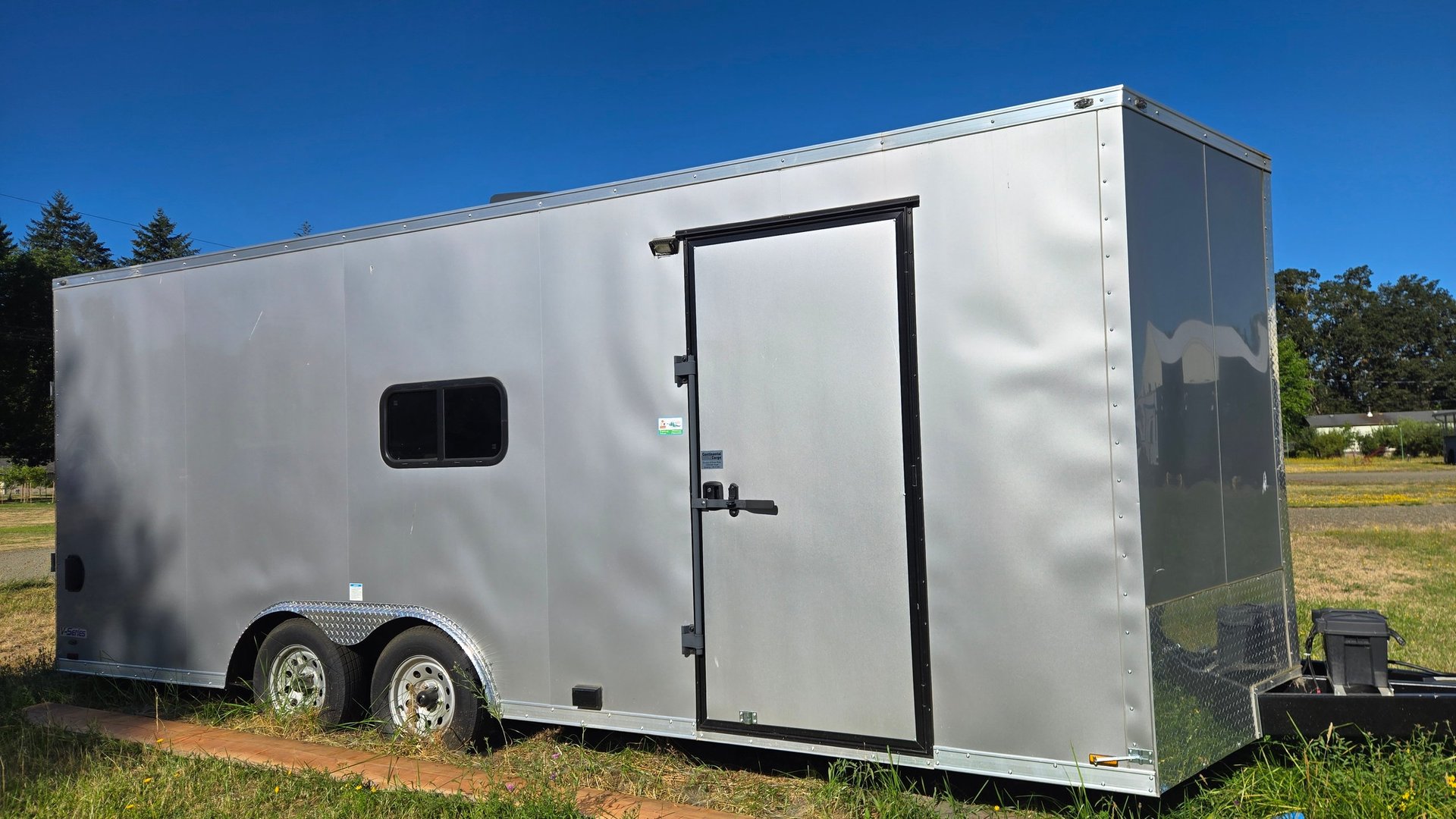 White enclosed cargo trailer with dual axles parked on grass field under clear blue sky