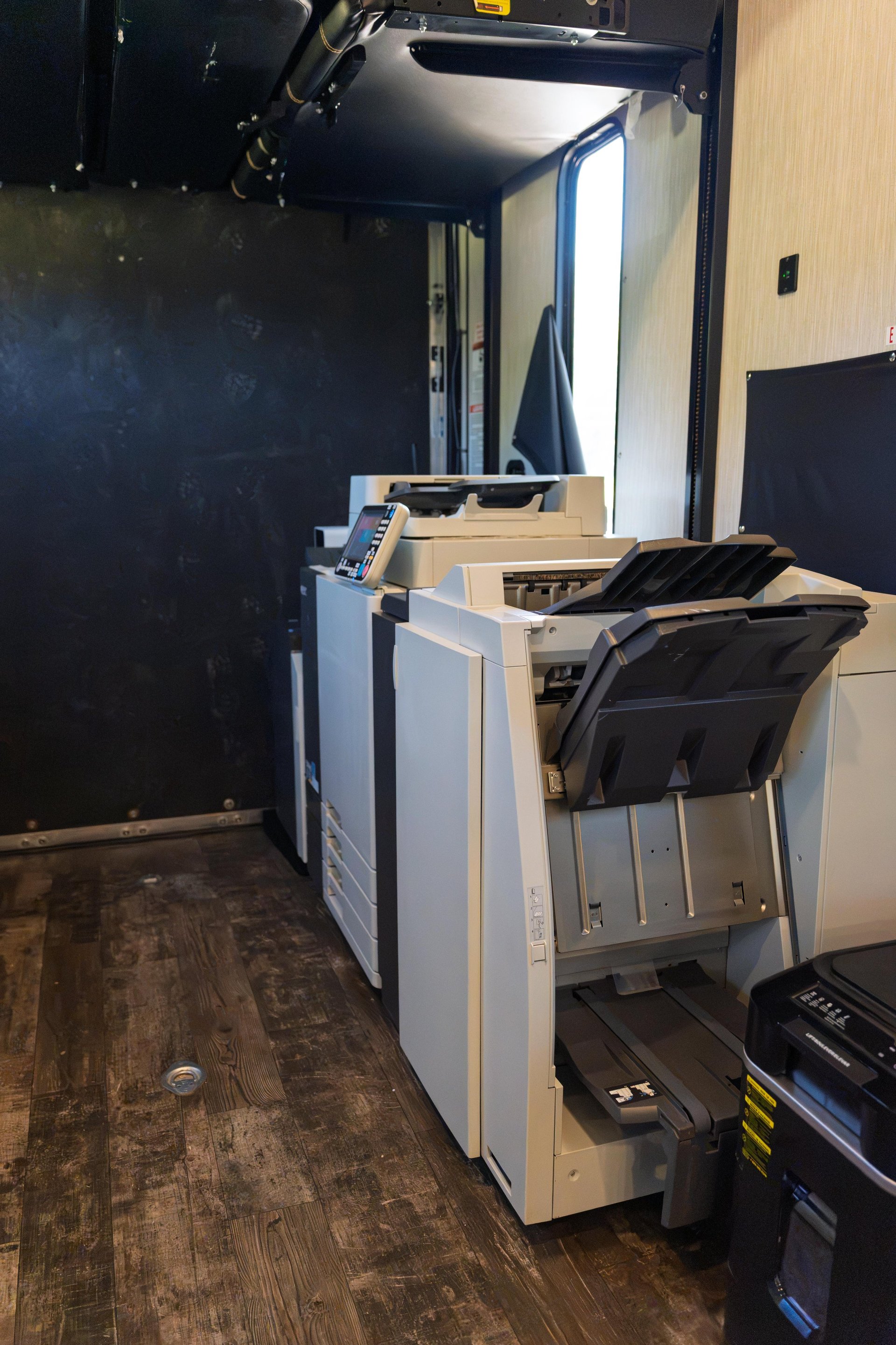 Commercial office copier and printer equipment with paper trays loaded with black materials in a small room with black walls and wood flooring