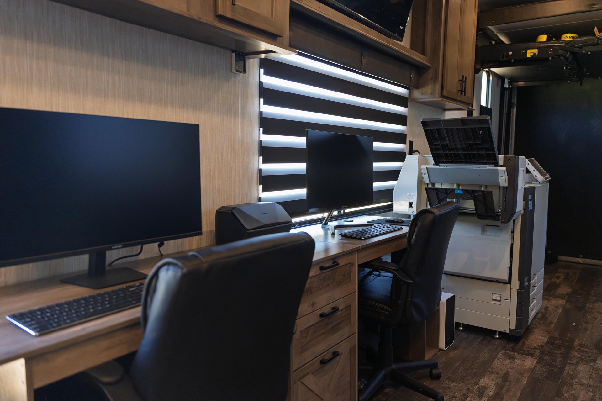 Modern office workspace with dual monitors, black office chairs, wooden desk, and printing equipment in a dark wood interior